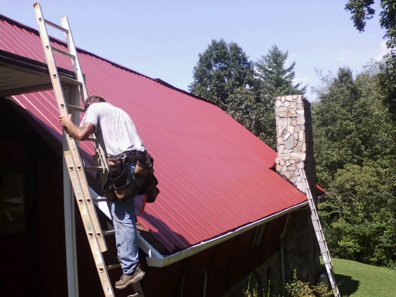 Roofer on a ladder beside a red metal roof and stone chimney — Soffit & Fascia Repair by Roofing Brothers in Guild